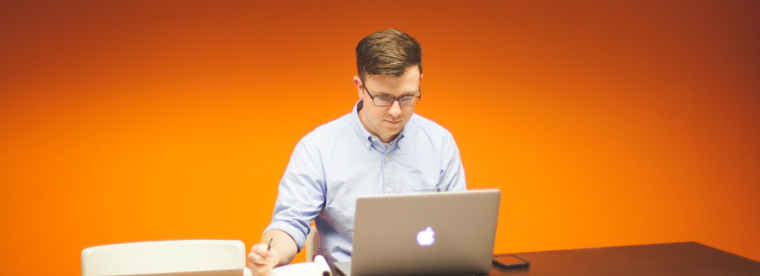 Young man in blue shirt working on laptop at desk against orange wall