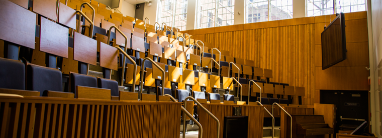 Tiered lecture hall with blue chairs and desk lamps with wood paneling and windows