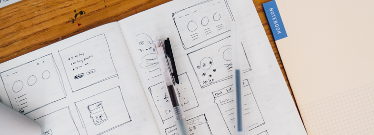 Overhead view of wireframe UI sketches on paper with pen and notebook on wooden table