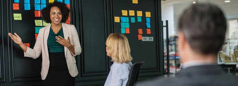 Woman in beige blazer presenting at workshop with colorful sticky notes on board