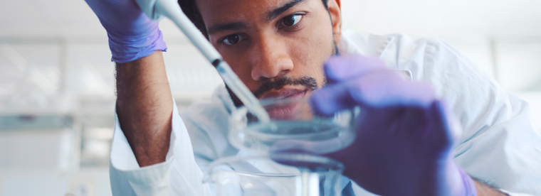 Laboratory researcher in white coat and blue gloves pipetting liquid into glass beaker